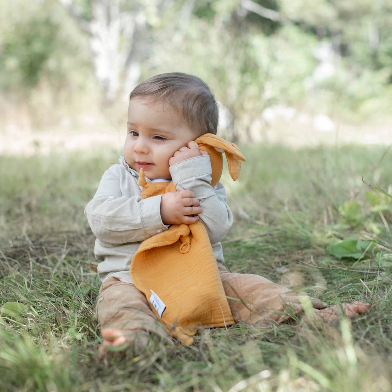 On voit un bébé assis sur l'herbe avec doudou lapin orange dans les bras.