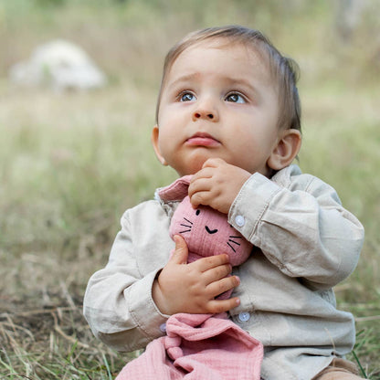 Nous voyons un bébé assis serrant dans ses bras un doudou en coton bio rose.