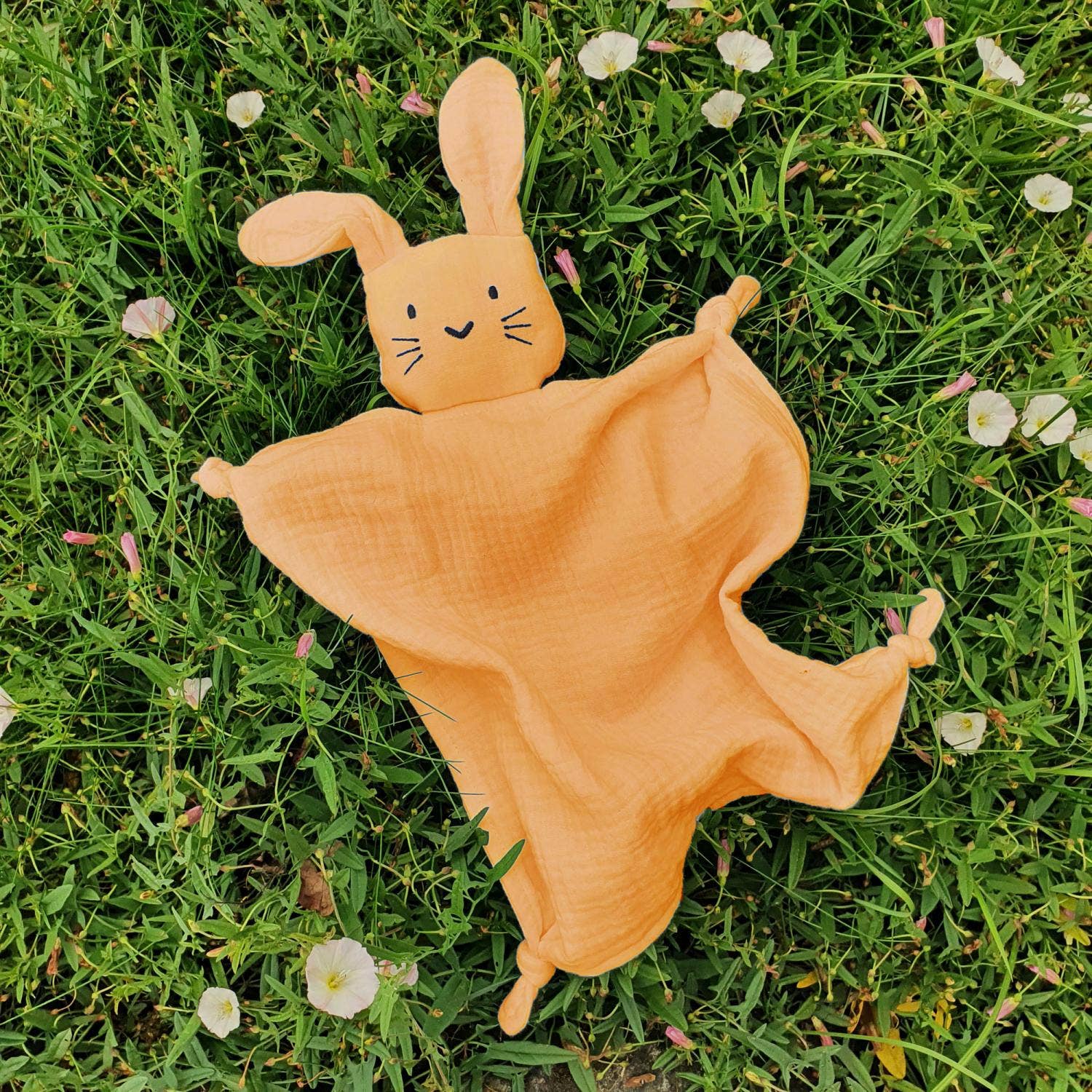 Doudou lapin en coton orange posé sur l'herbe verte entouré de fleurs de printemps.