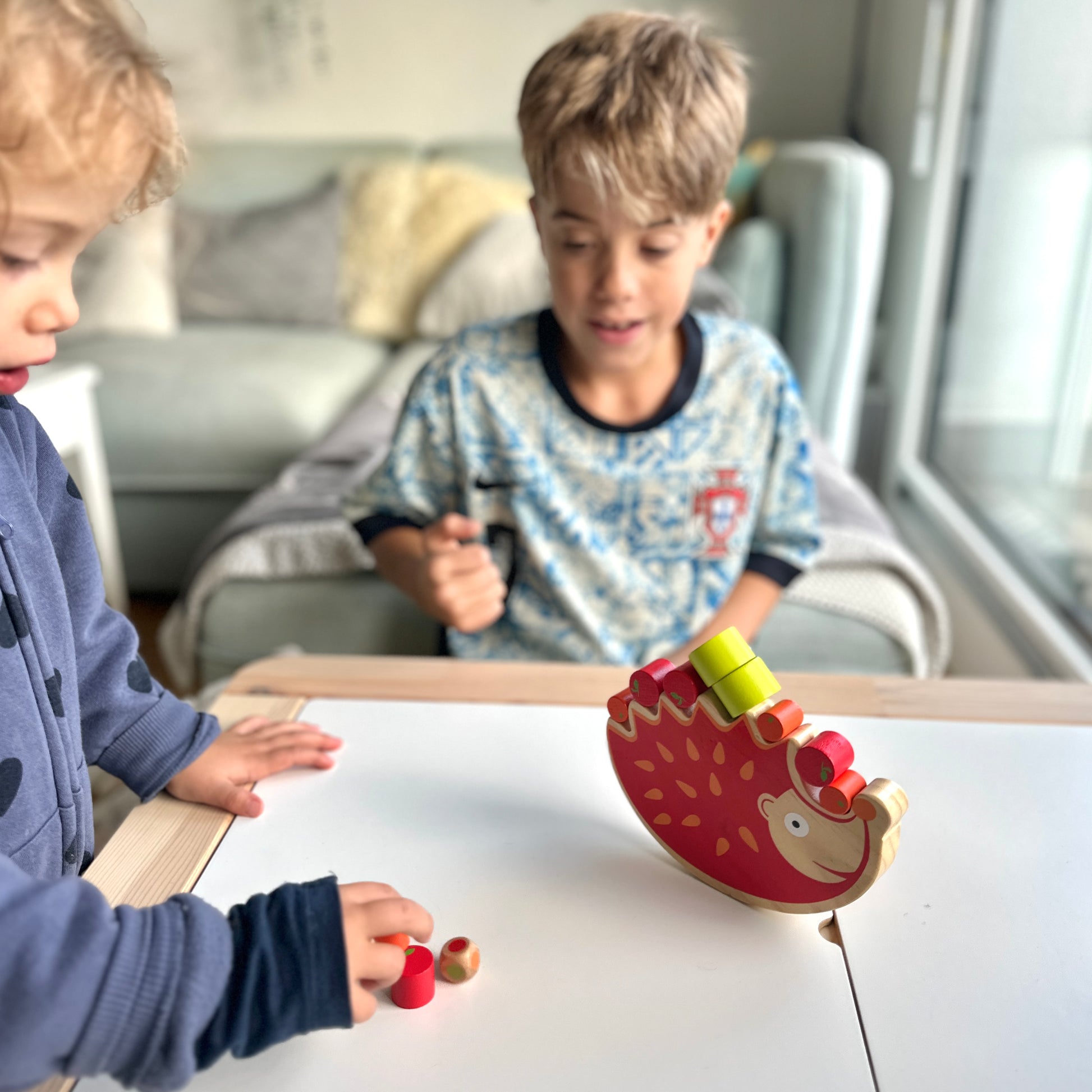 On voit une partie du jeu du balancier en cours entre une petite fille et son grand frère. Le hérisson est posé au centre de la table et avec des pièces sur le dos. La petite fille s'apprête à poser une pièce sur le dos du hérisson.