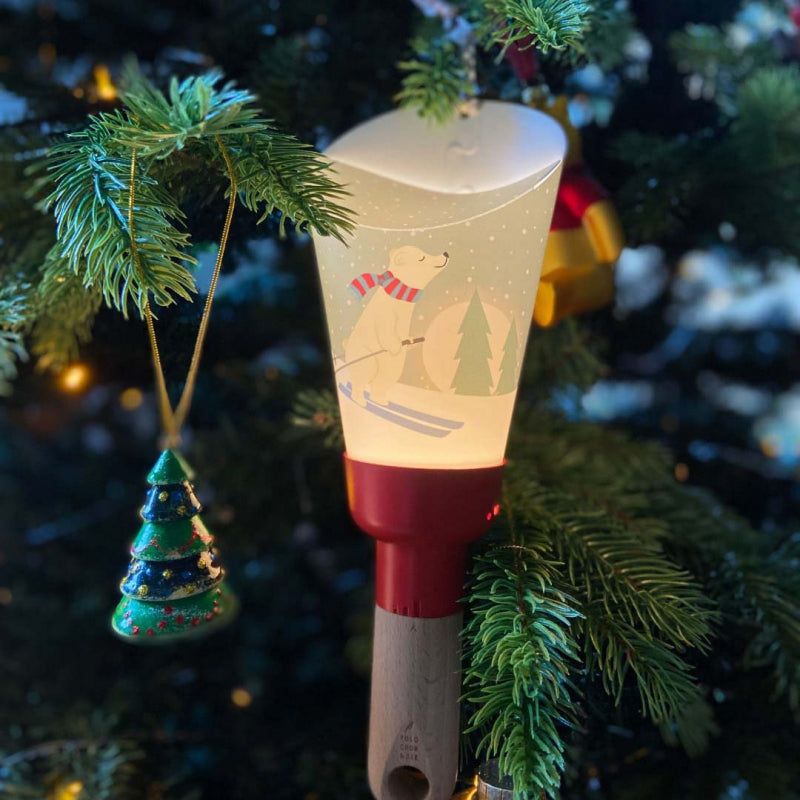 la lampe nomade ours skieur est suspendue allumée au sapin de noël à coté d'autres décorations de noël.